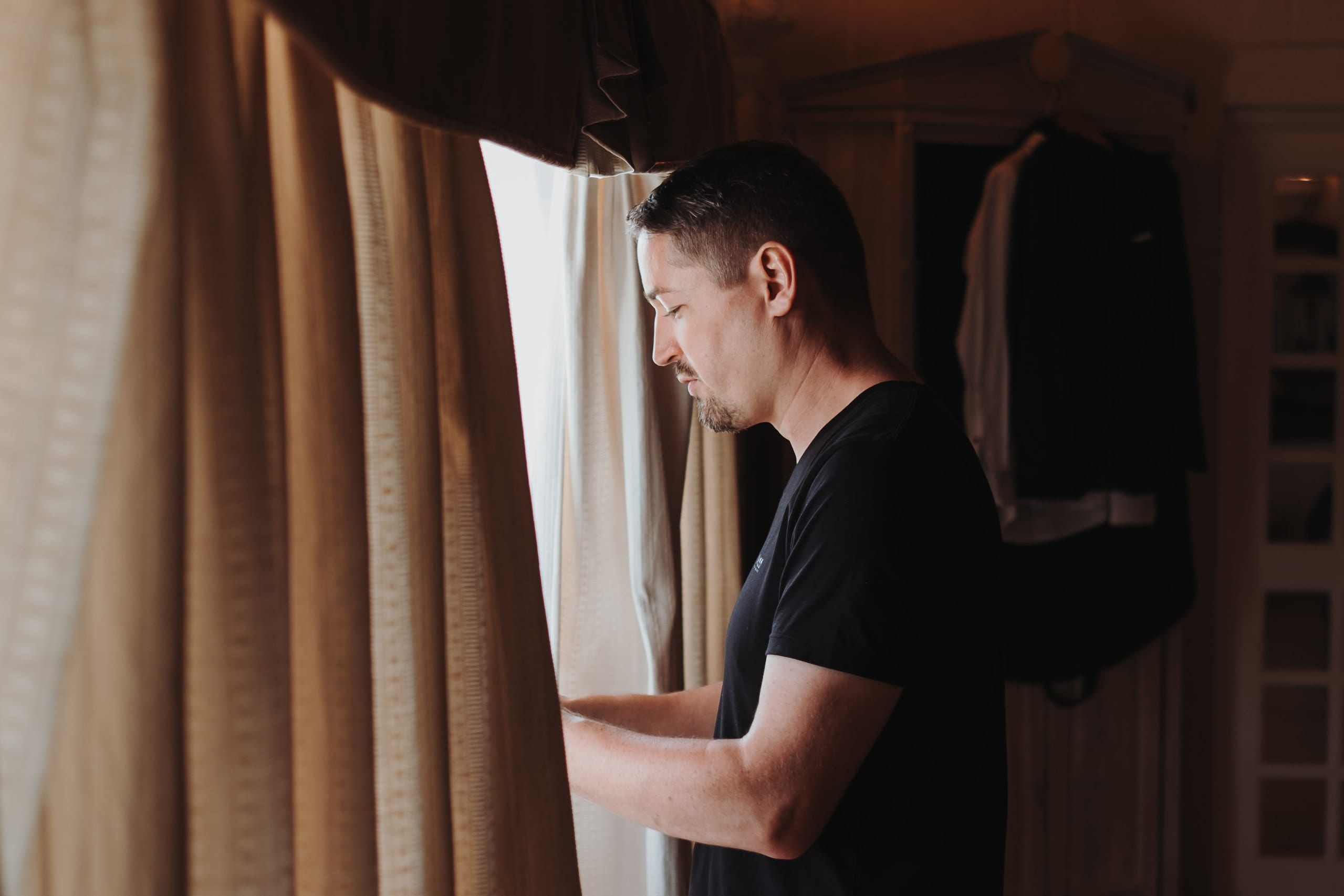 Der Bräutigam im schwarzen T-Shirt am Fenster, hinter ihm hängt sein Anzug im Schrank