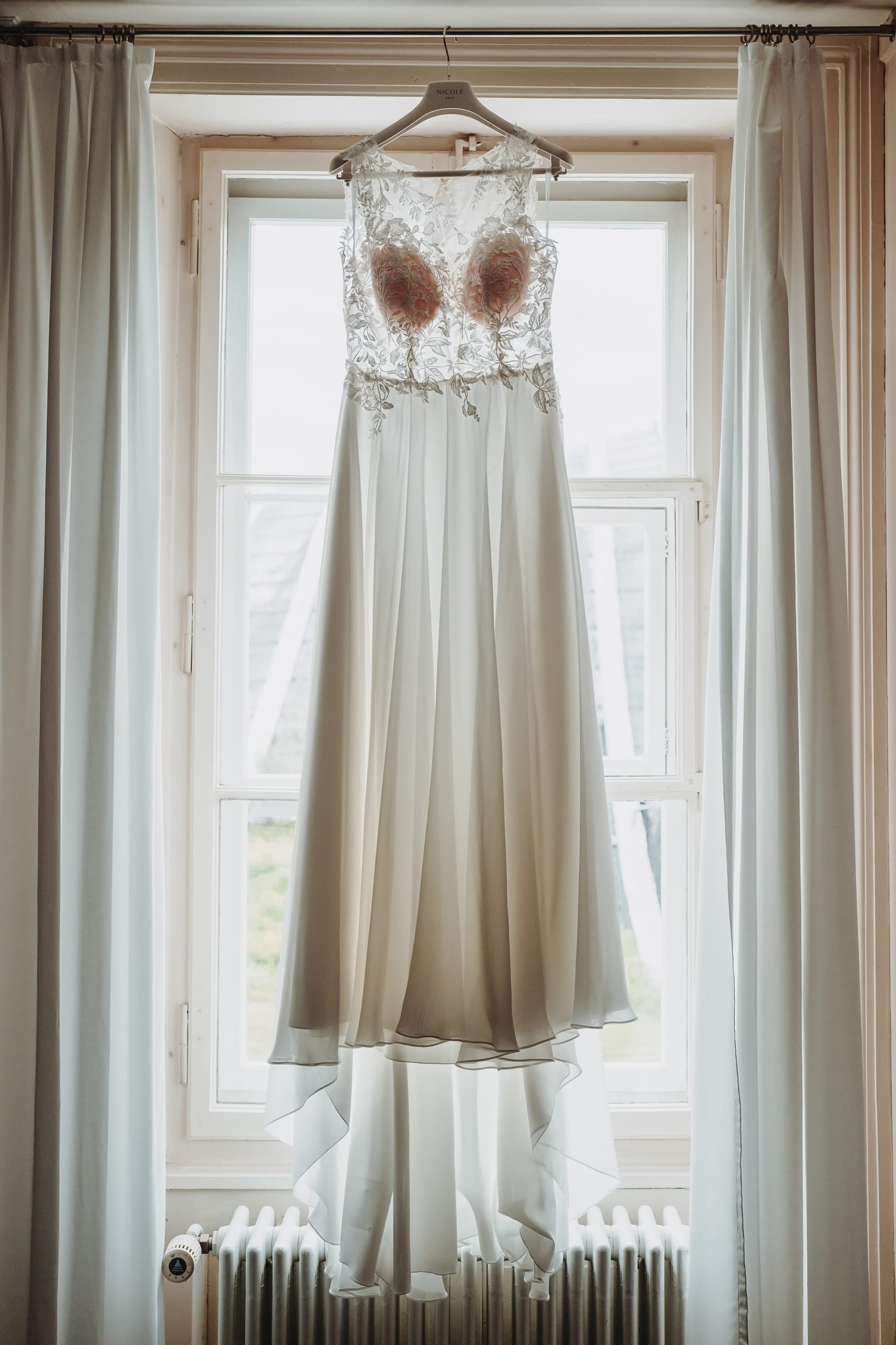 Das Brautkleid hängt vor einem hohen Fenster