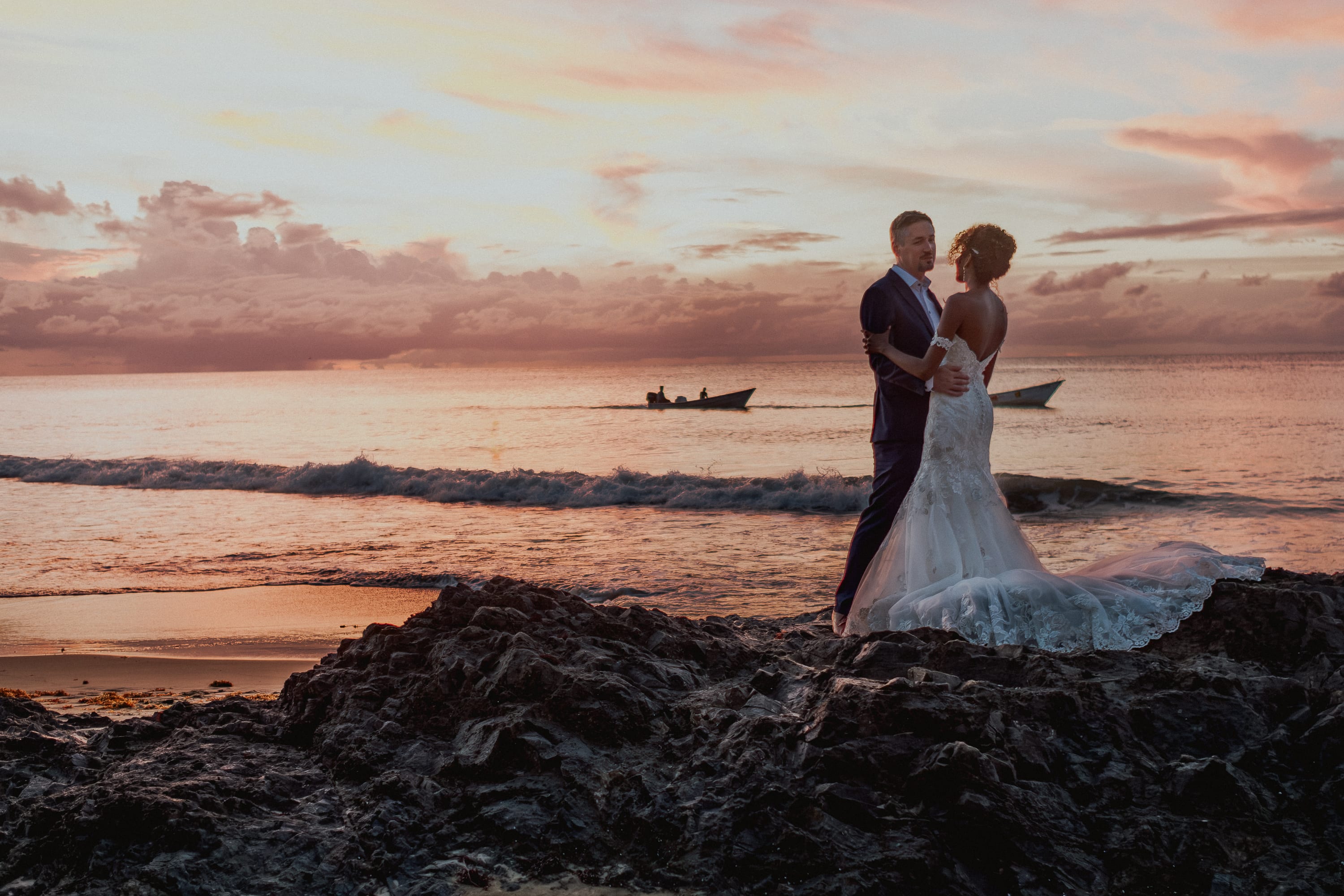 Das Brautpaar auf Felsen am Strand von Tobago vor rosa Abendhimmel, auf dem Meer zwei kleine Boote