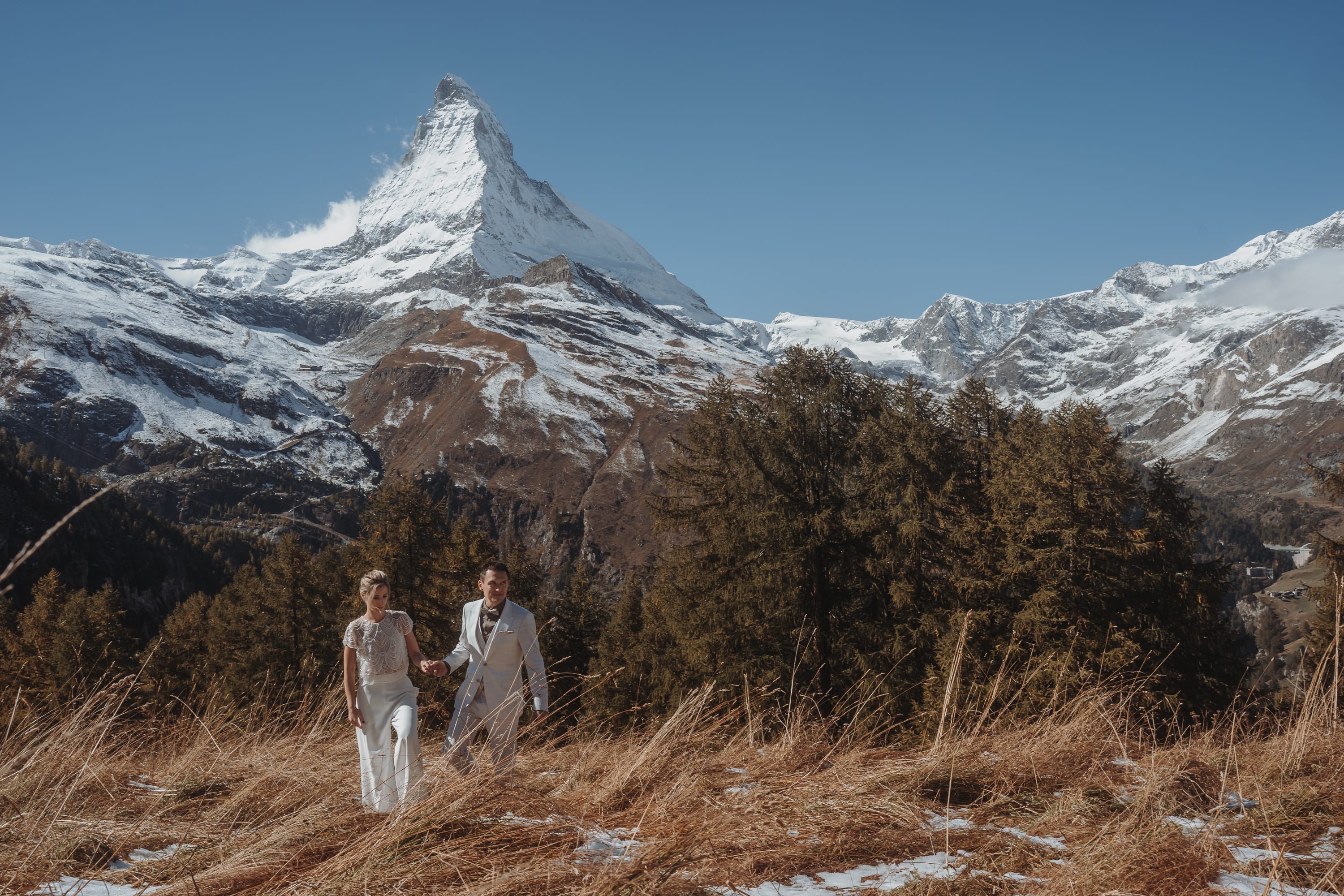 Das Brautpaar geht Hand in Hand durch das goldene Gras, darüber das Matterhorn