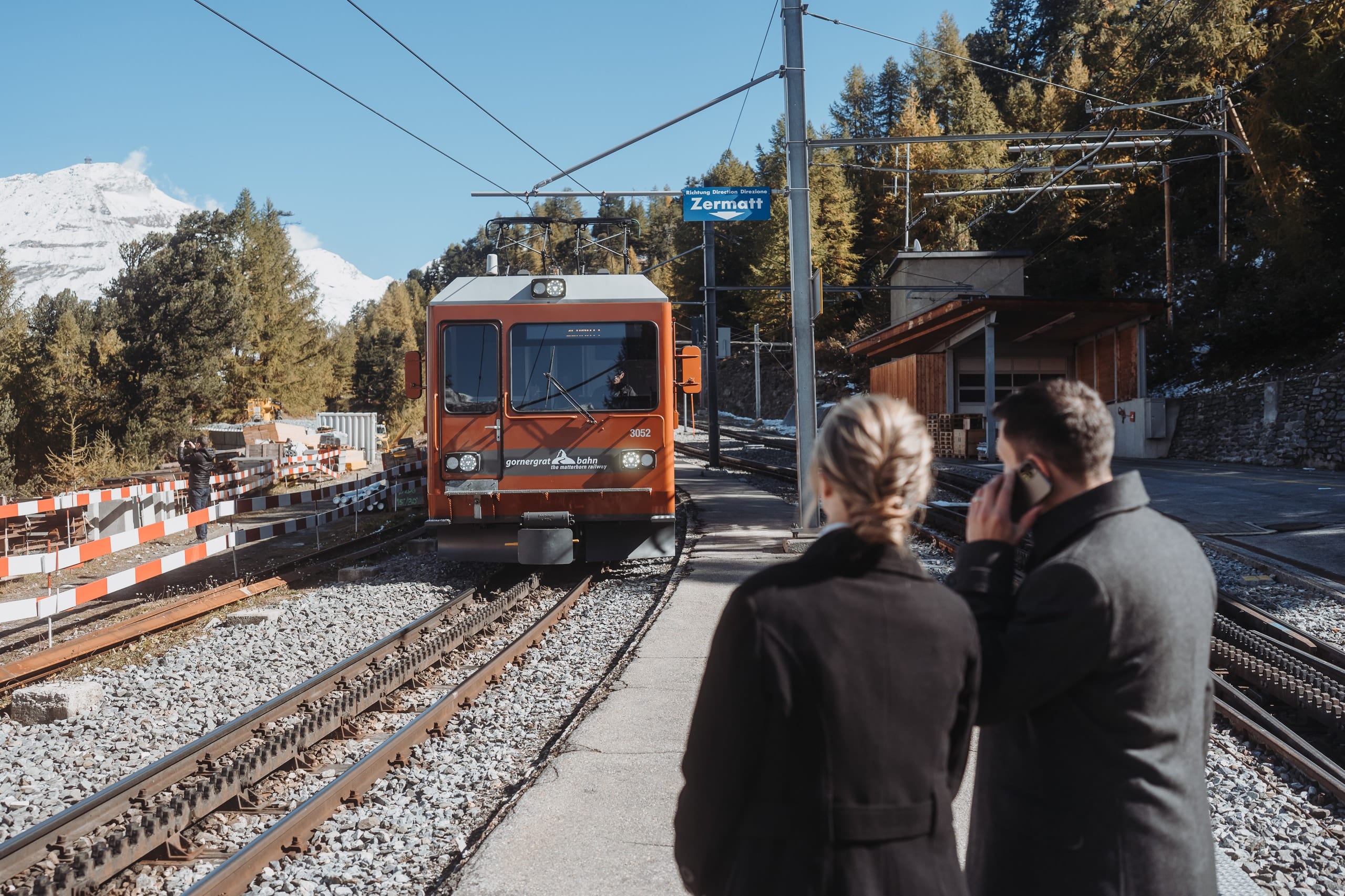 Das Brautpaar in Mänteln am Bahnsteig in Zermatt, die rote Gornergratbahn fährt ein