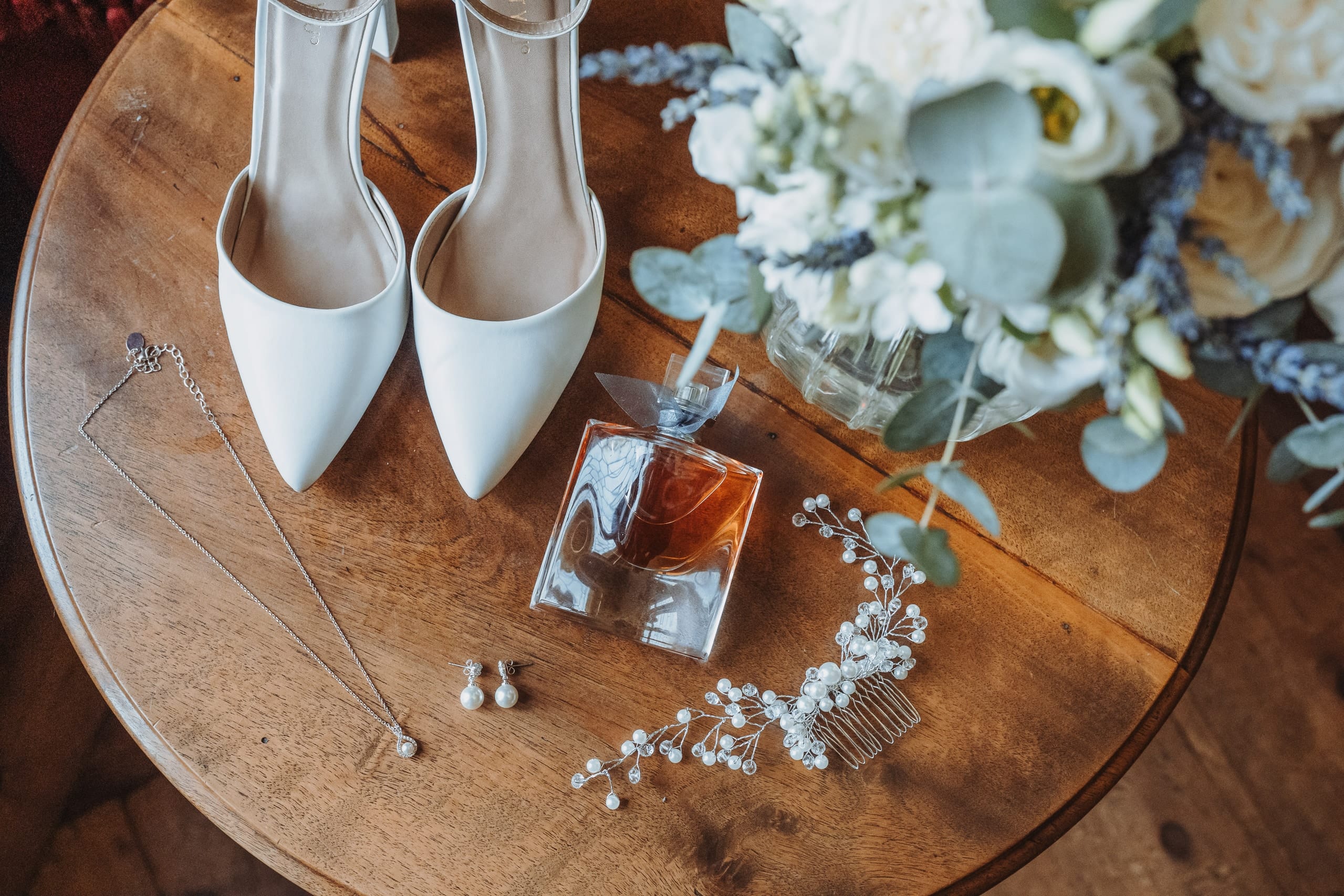 Brautschuhe, Parfum, Schmuck und Blumen auf einem runden Holztisch
