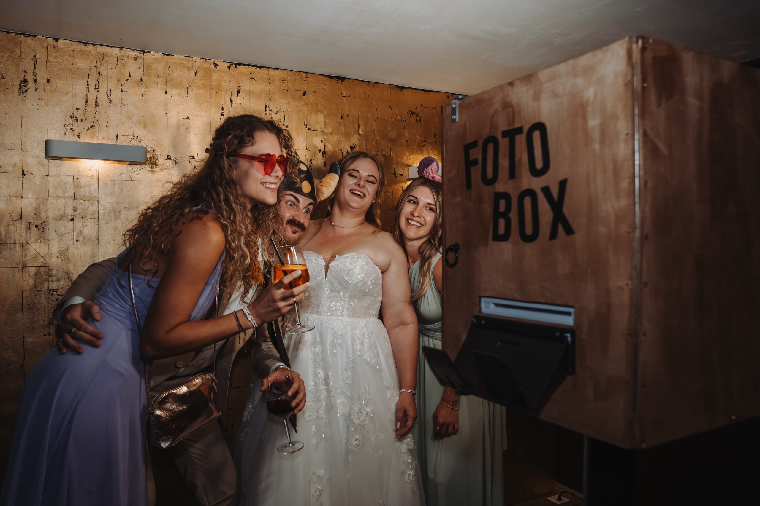 Fotobox an einer Hochzeit: Die Braut und ihre Gäste rücken mit Hüten und Brillen vor der Fotobox zusammen