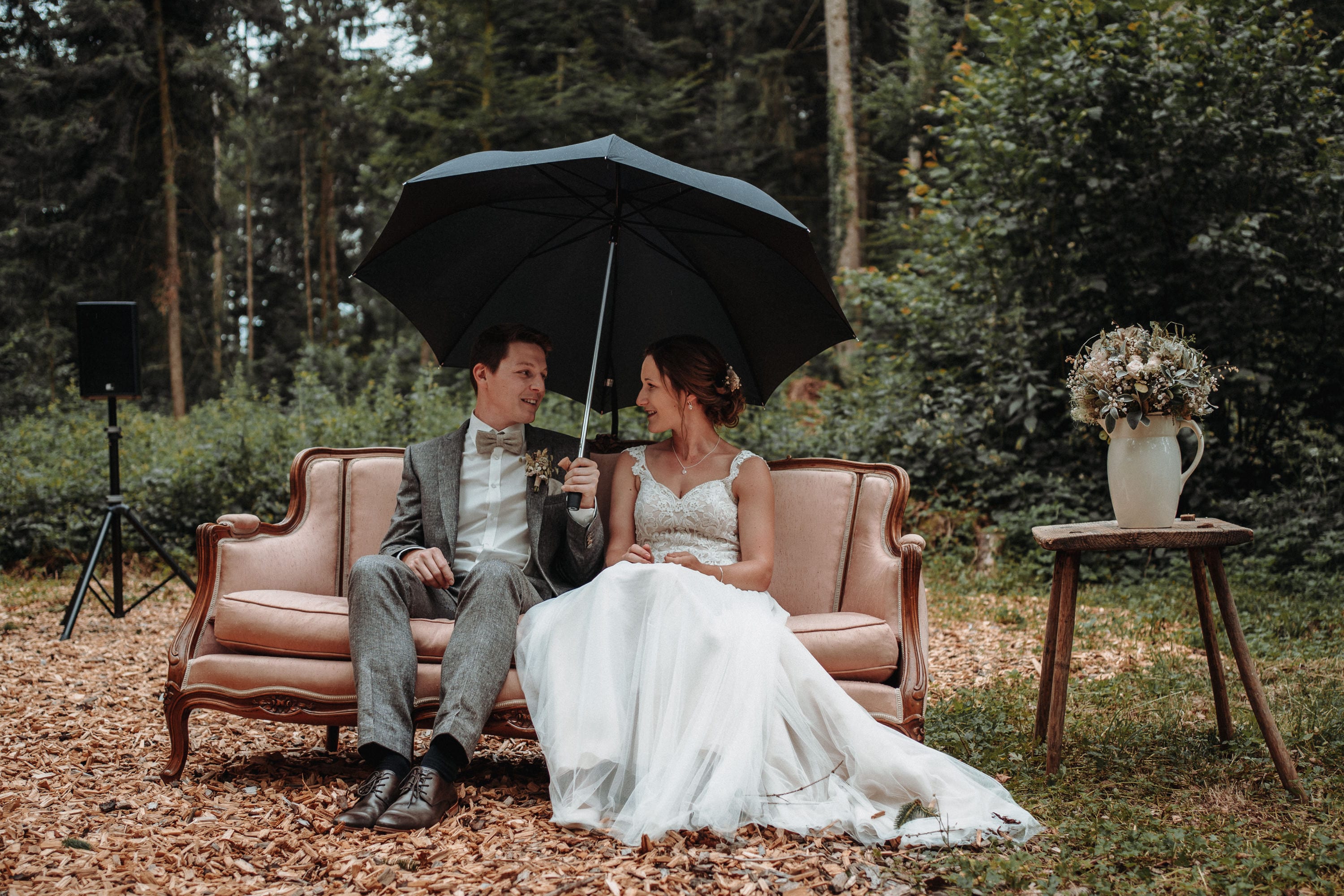 Das Brautpaar sitzt bei der freien Trauung im Wald unter einem Regenschirm auf einem rosa Sofa