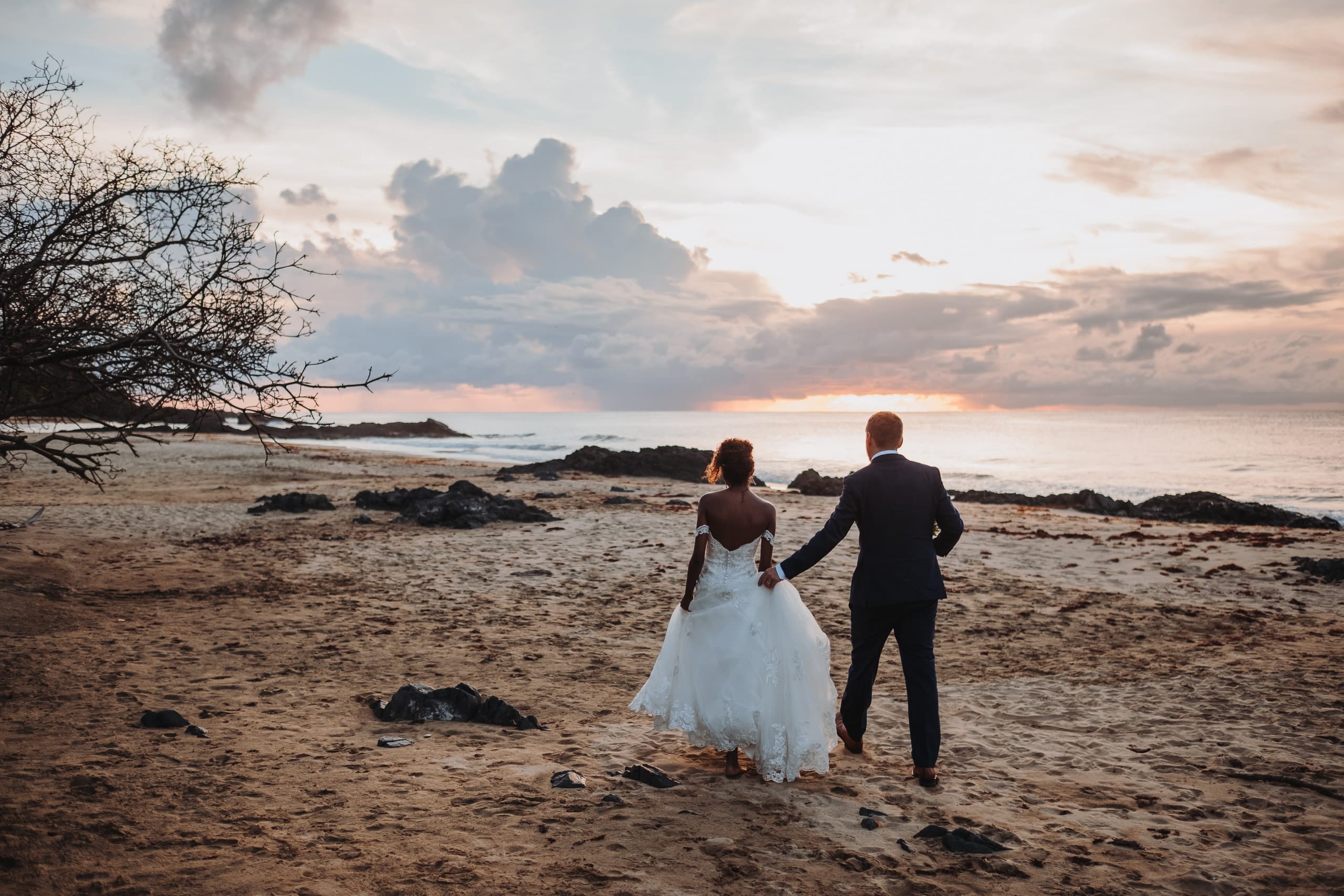 Das Brautpaar geht Hand in Hand über den Strand von Tobago, am Horizont scheint die Sonne durch eine Wolkenlücke