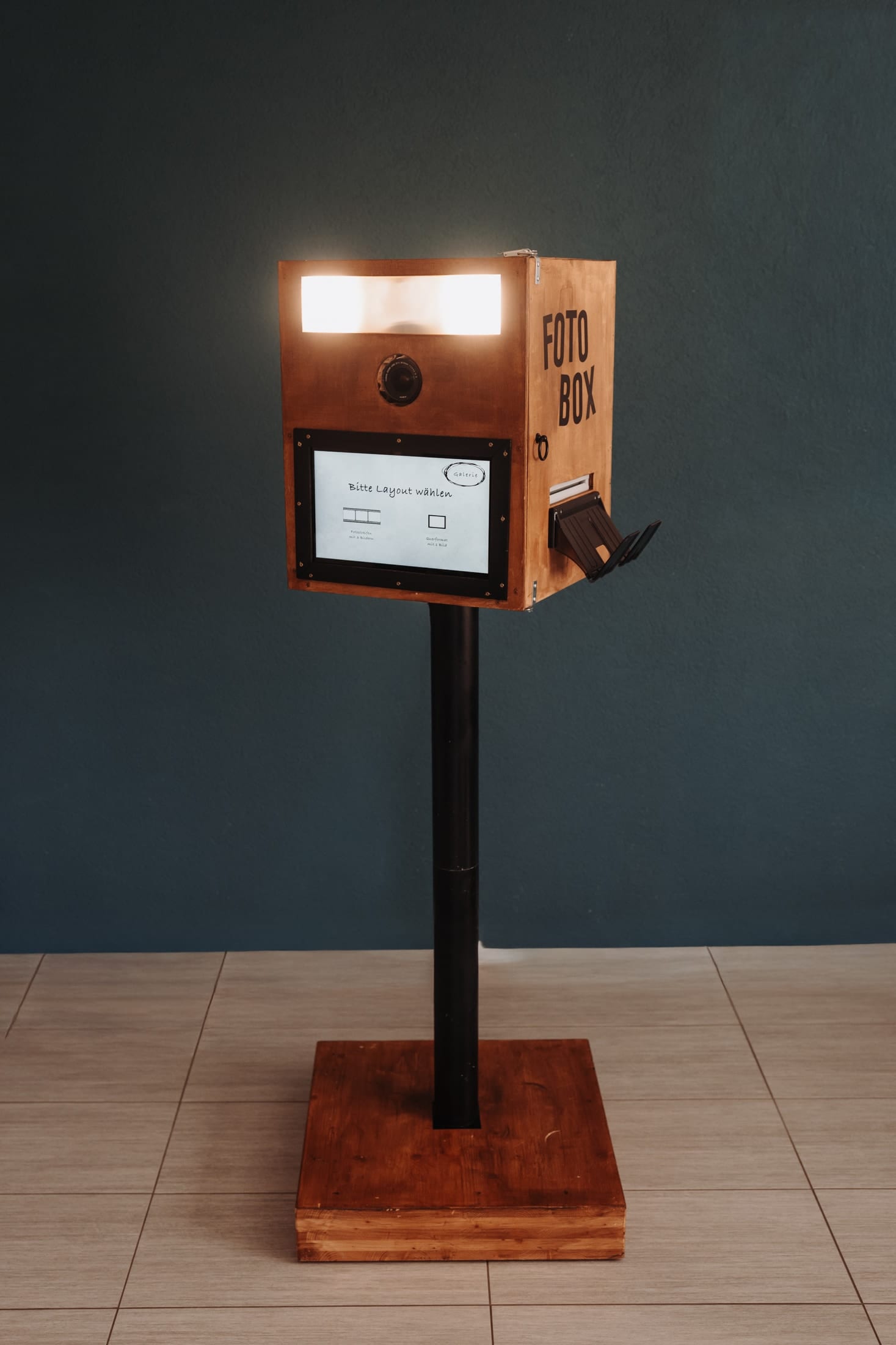 Die Fotobox aus Holz auf ihrem schwarzen Standfuss mit Holzsockel, oben leuchtet der Blitz