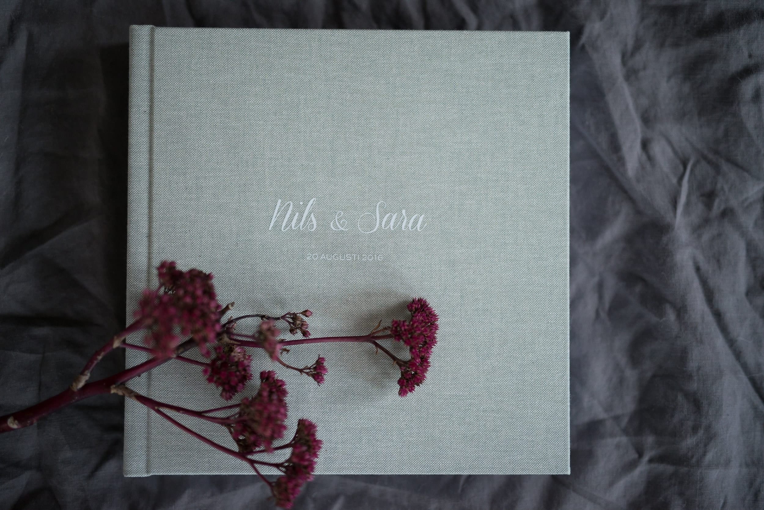 Hochzeitsalbum mit hellgrauem Leineneinband und weisser Prägung auf dunklem Stoff, darüber ein Zweig mit dunkelroten Trockenblumen