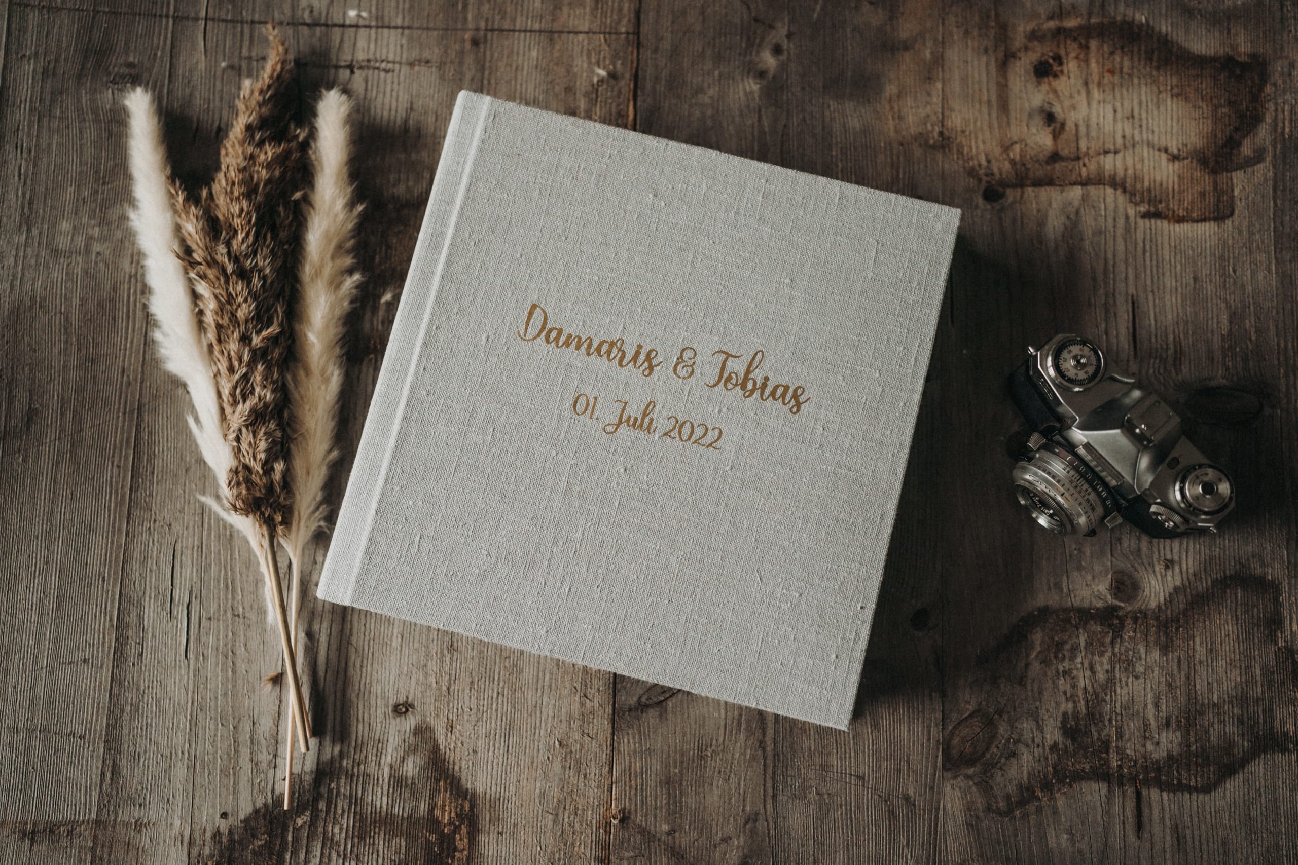 Hochzeitsalbum mit hellem Leineneinband und Namensprägung auf einem Holztisch, daneben Trockengräser und eine alte Kamera