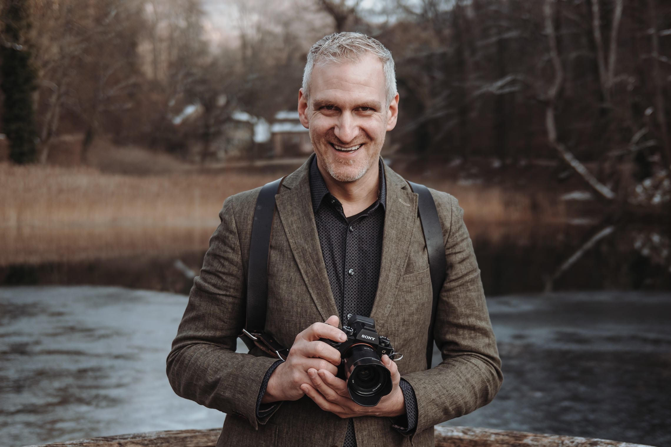 Claudio Montefusco steht lächelnd mit seiner Kamera an einem Bach, im Hintergrund Bäume und ein Haus
