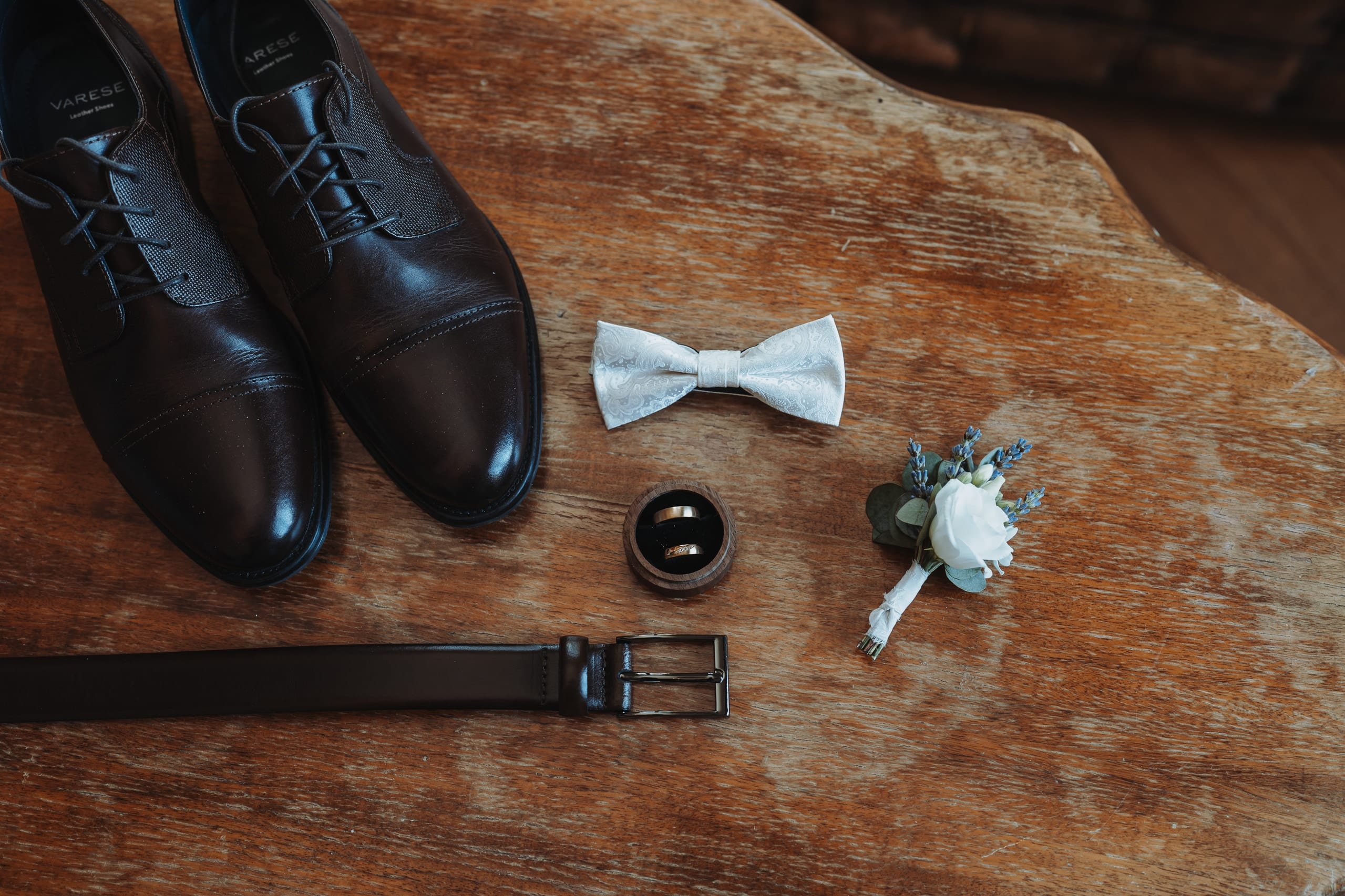 Schuhe, Fliege, Gürtel, Eheringe und Ansteckblume des Bräutigams auf einem Holztisch