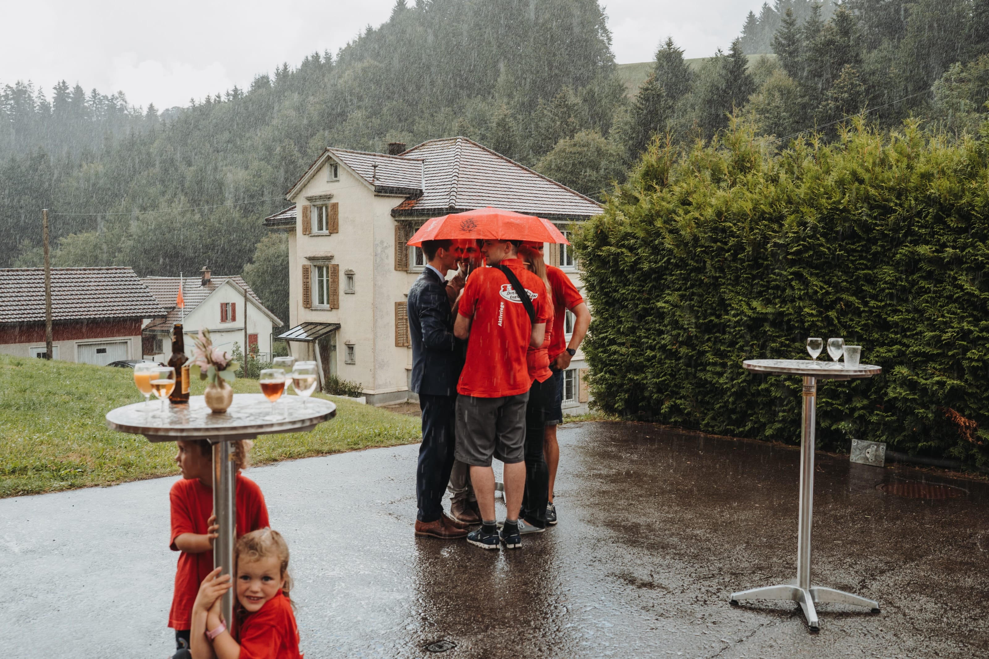 Apéro im Regen: Gäste unter einem roten Schirm vor einem alten Haus
