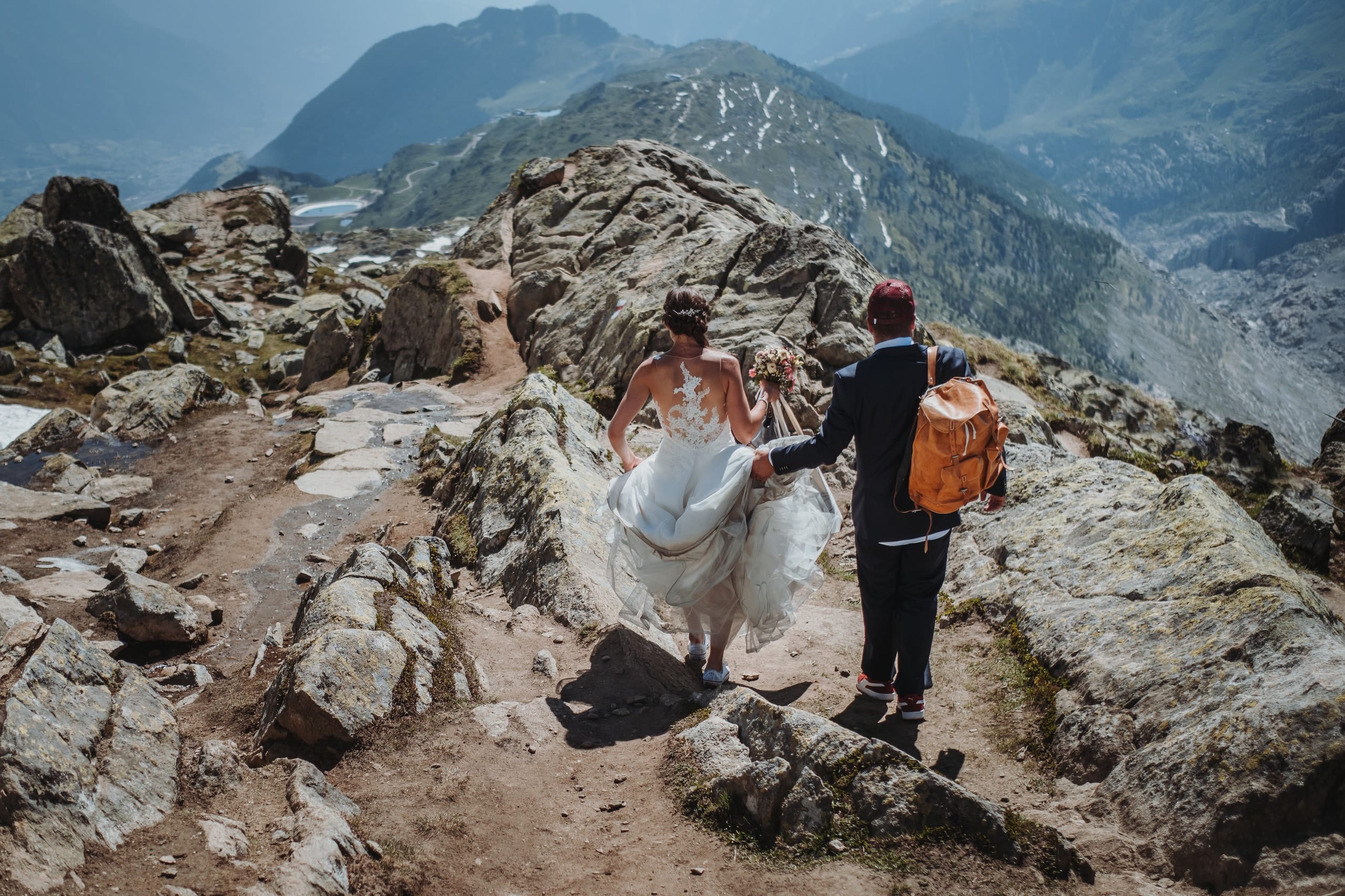 Braut und Bräutigam steigen Hand in Hand über einen felsigen Bergweg bei der Bettmeralp, er mit Rucksack