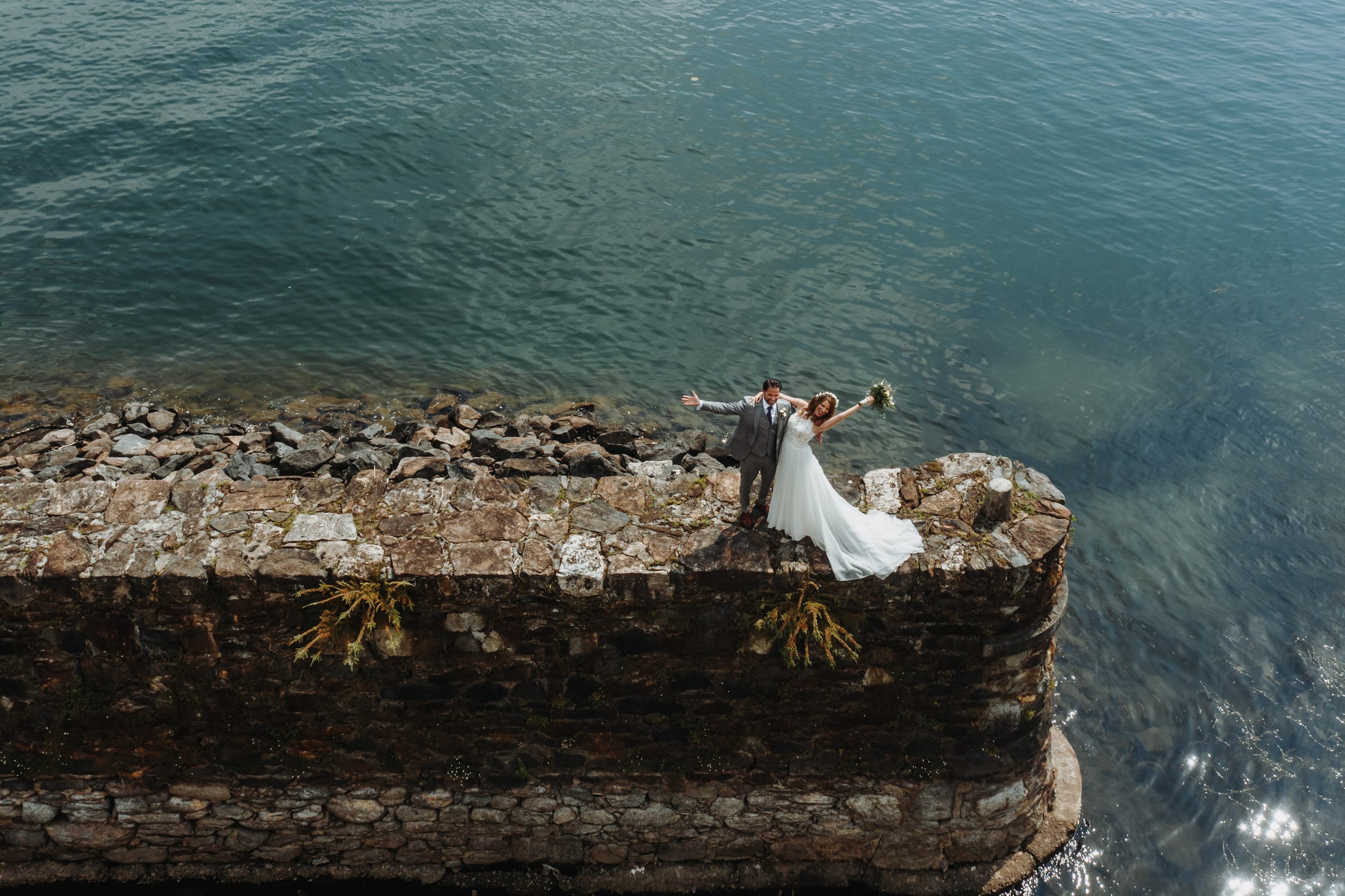Das Brautpaar auf einer Steinmauer am Wasser, von oben fotografiert