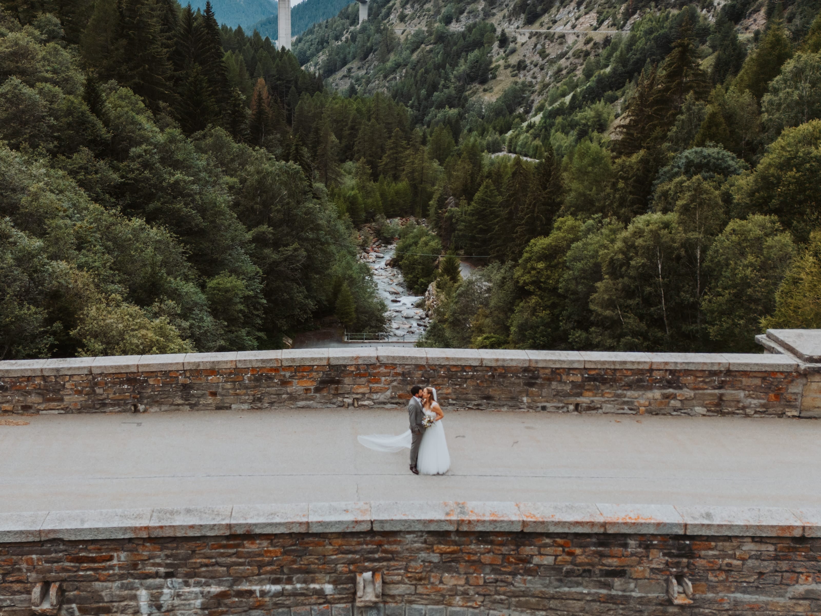 Das Brautpaar umarmt sich am Simplon auf einer Steinbrücke über einer bewaldeten Schlucht, von oben gesehen
