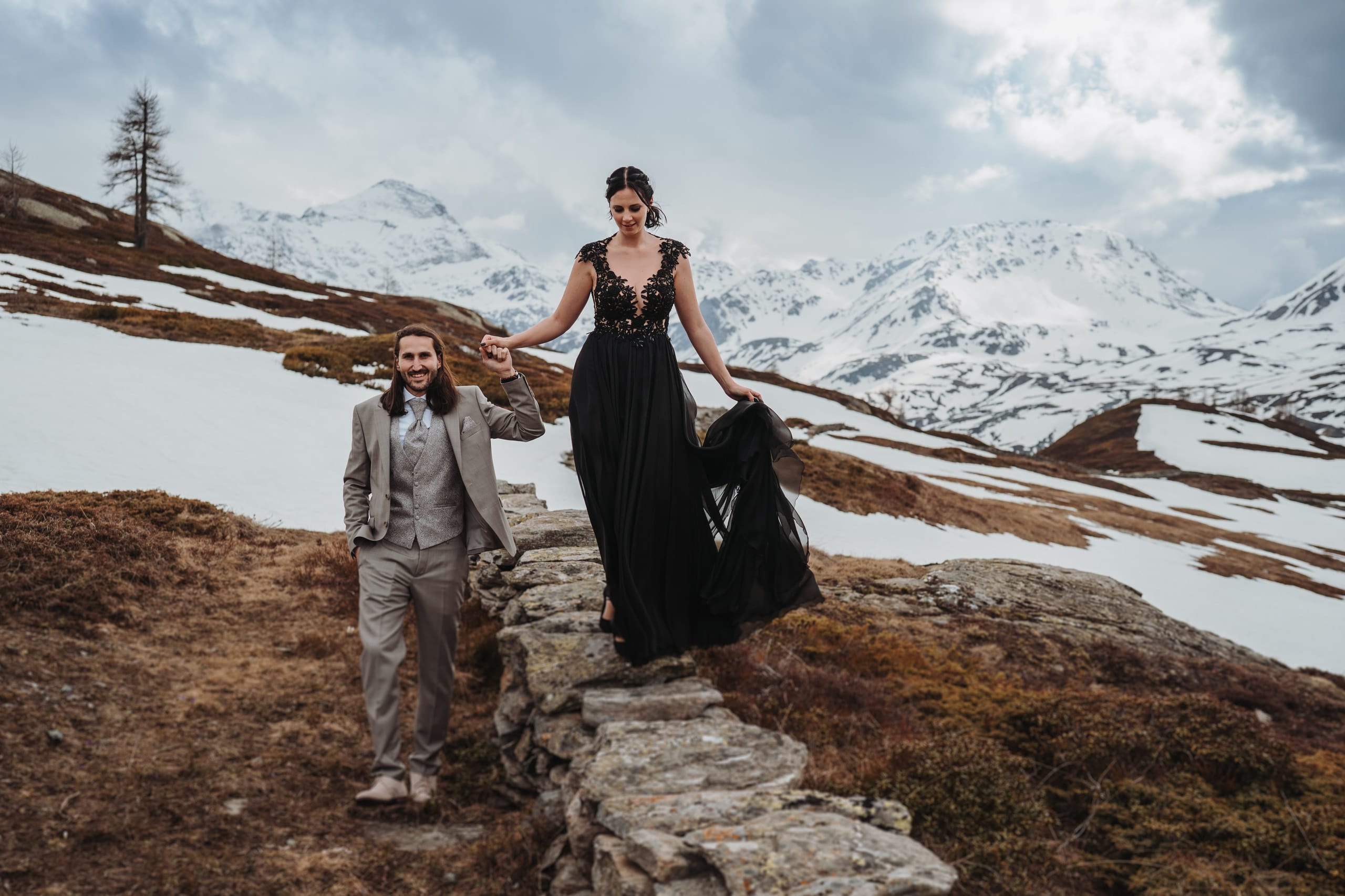 Die Braut im schwarzen Kleid balanciert am Simplon über eine Steinmauer, der Bräutigam hält ihre Hand