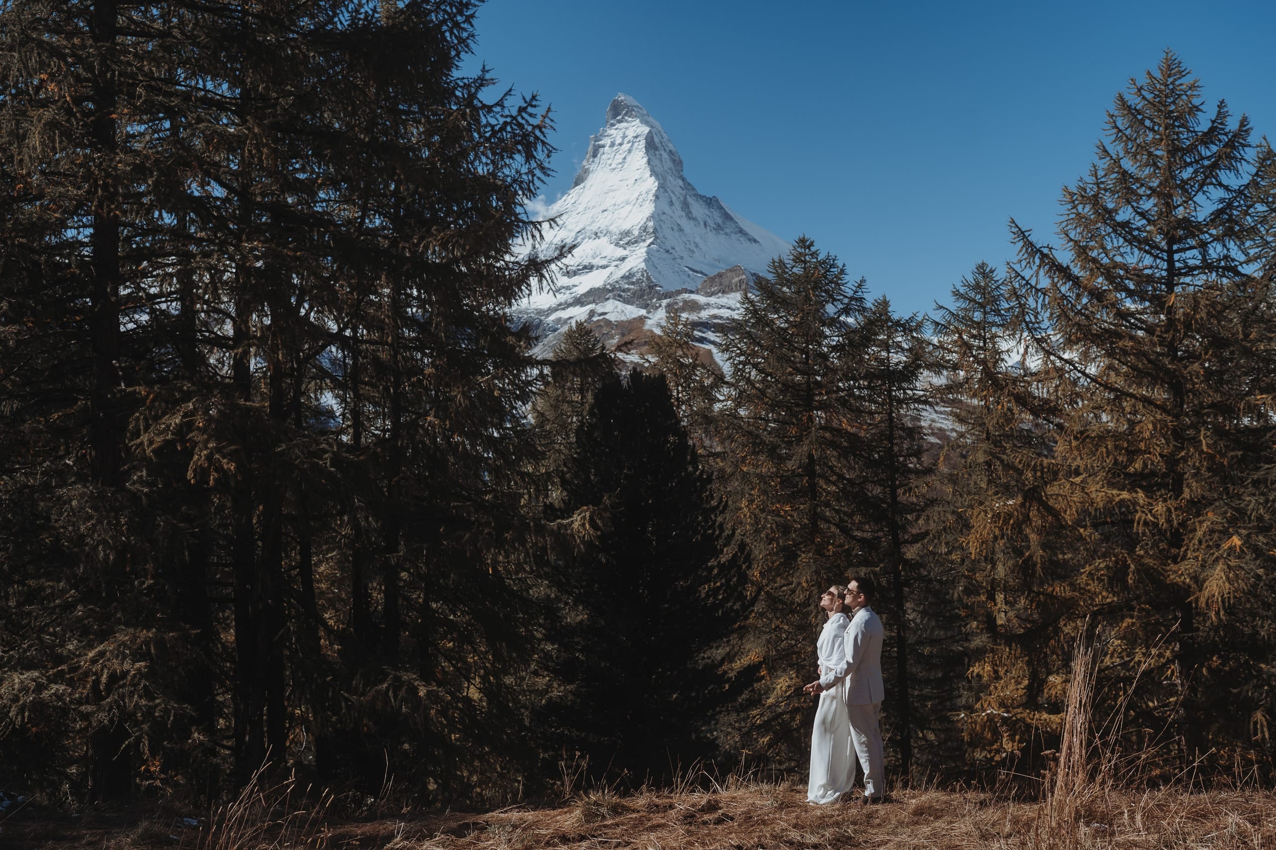 Das Brautpaar klein zwischen hohen Bäumen bei Zermatt, in der Lücke das Matterhorn