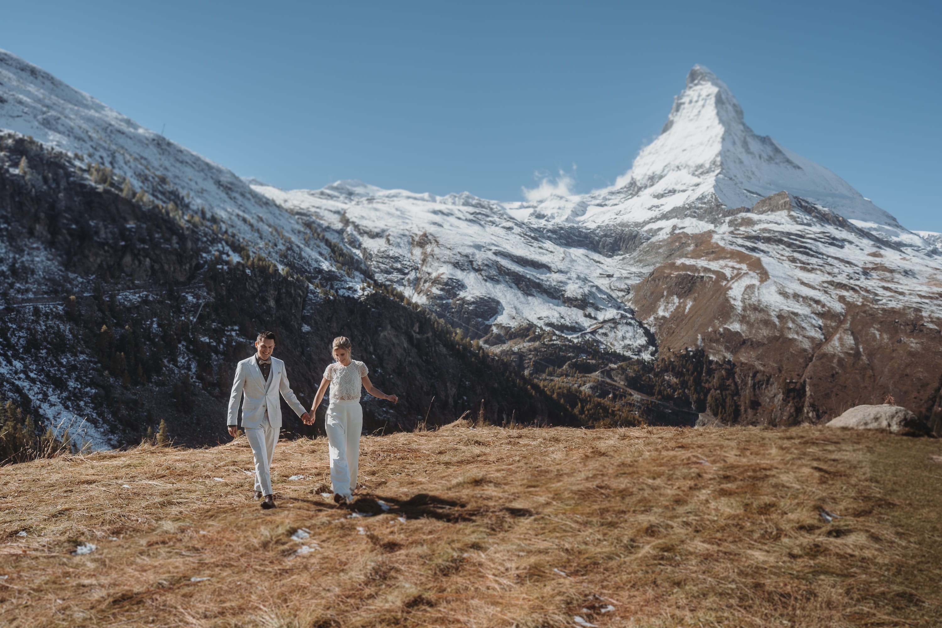 Das Brautpaar geht an seinem Hochzeitstag in Zermatt Hand in Hand über eine Bergwiese, dahinter das Matterhorn